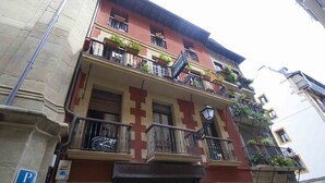 A multi-story building with a red and beige facade, balconies with plants, and a visible 'P' parking sign. - Coeur | The Rentals Collection (San Sebastián)