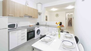 A compact kitchen with a microwave, oven, and washing machine. There is a dining table set with plates, glasses, and a vase with flowers. - Coeur | The Rentals Collection (San Sebastián)