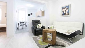A modern living room with a black leather chair, a white sofa, a wooden side table, and a dining area with a table and chairs. - Coeur | The Rentals Collection (San Sebastián)