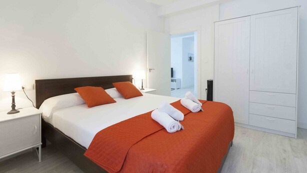 A hotel room with a bed, white nightstands, a wardrobe, and a door leading to another room. - Coeur | The Rentals Collection (San Sebastián)