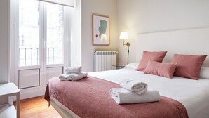 A hotel room with a bed, a bedside table, a lamp, a radiator, and a window with a view of buildings. - Hondarribia | The Rentals Collection (San Sebastián/Donostia)