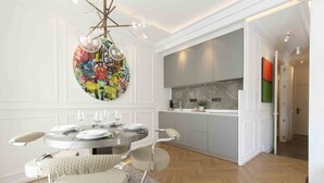 A dining area with a round table, chairs, and a chandelier. There is a kitchen in the background with cabinets and a countertop.