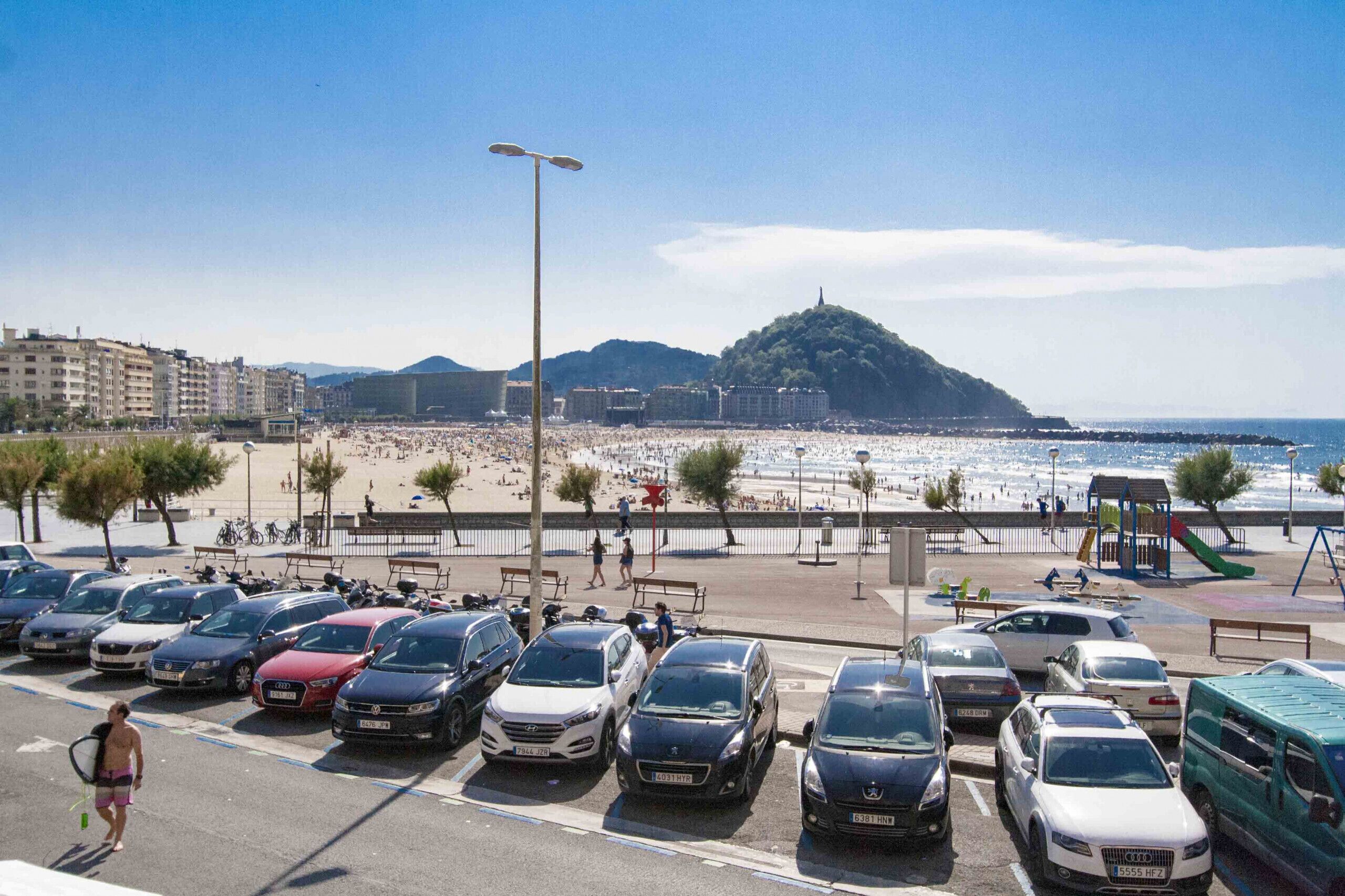 Een parkeerplaats met auto's, een strand en een heuvel op de achtergrond.
