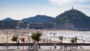 A crowded beach with people sunbathing and swimming, a large hill in the background, and buildings along the shore.