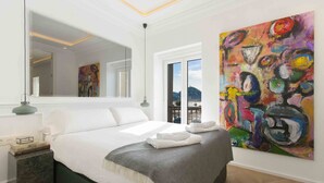 A modern bedroom with a large bed, a mirror, a painting, and a view of the city.