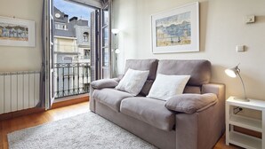 A living room with a grey sofa, two white cushions, a grey rug, a floor lamp, a small side table, and two framed artworks on the wall.
