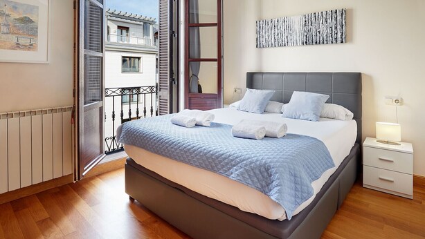 A bedroom with a bed, a nightstand, a bedside table, a window with a view of buildings, and a painting on the wall.