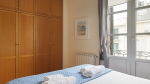 A bedroom with a large wooden wardrobe, a bed with a blue quilt, and a window with a view of buildings.