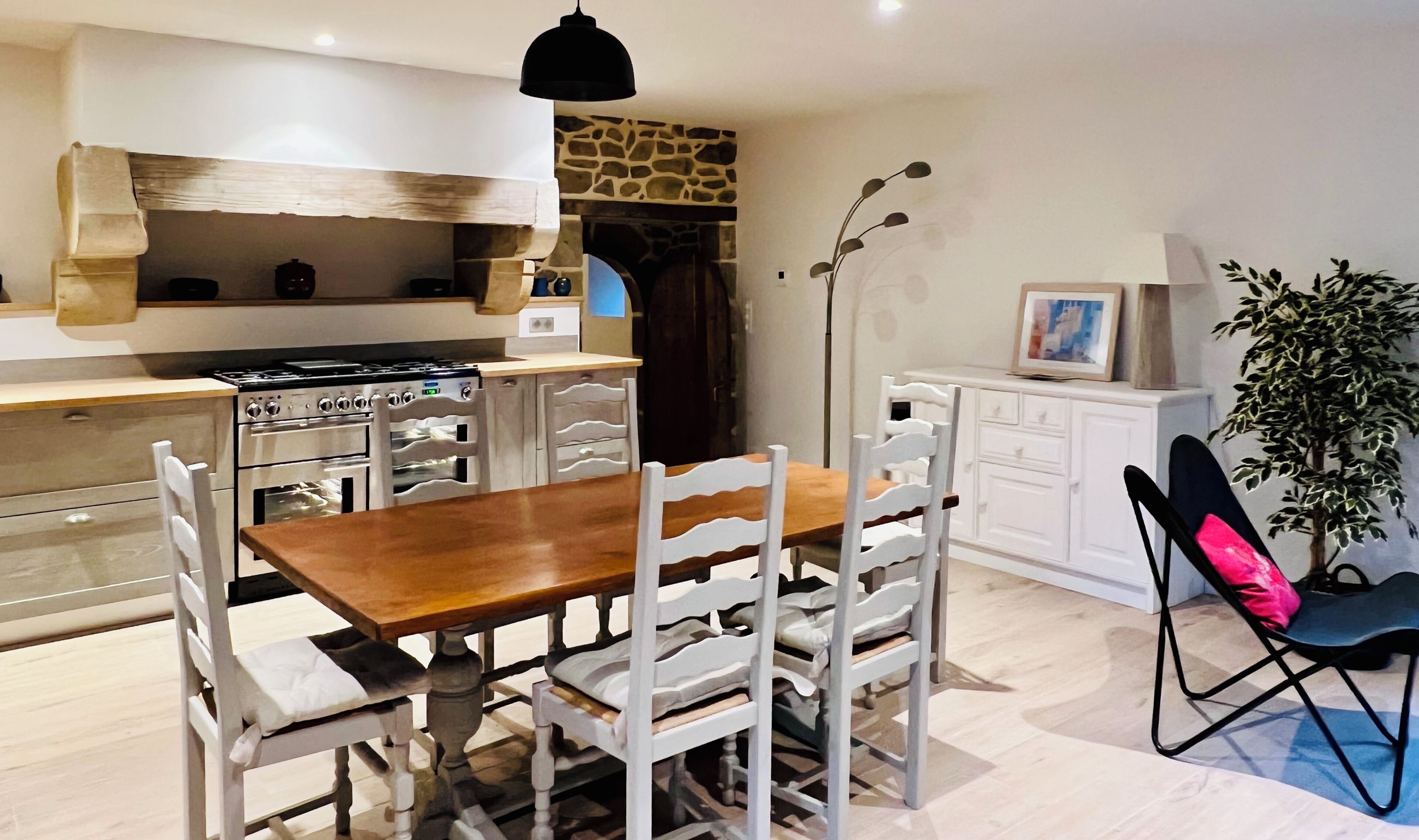 A modern kitchen with a wooden table and white chairs, a stone fireplace, and a dining area with a white cabinet and a plant.