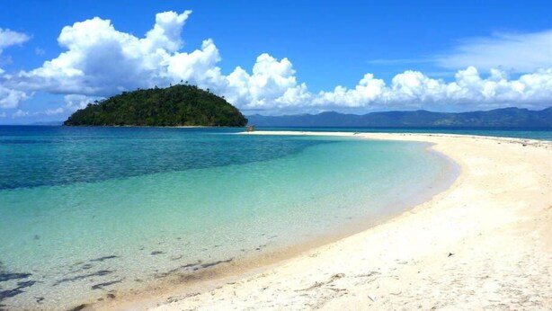Une plage à l’eau turquoise et limpide, un rivage sablonneux et une petite île couverte d’une végétation luxuriante.