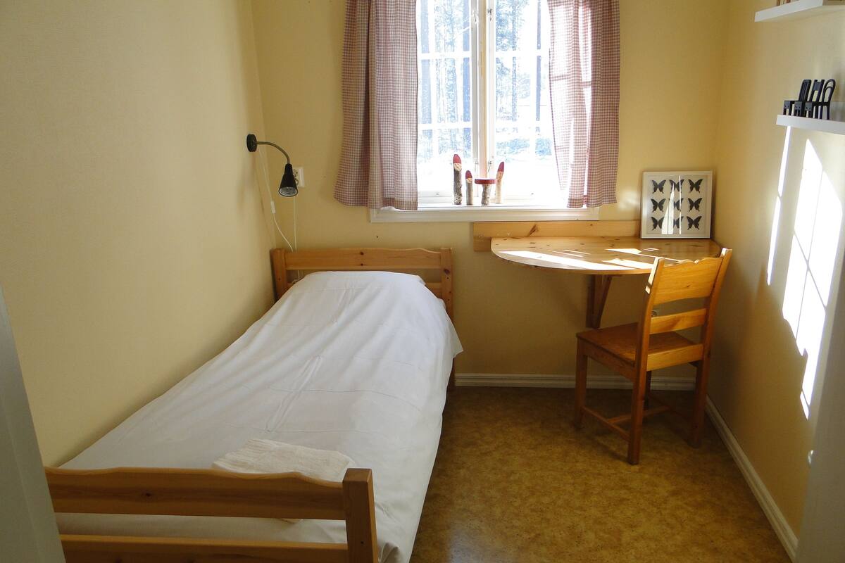 Una habitación pequeña y acogedora con una cama individual, una silla de madera, una mesita y una ventana con cortinas.
