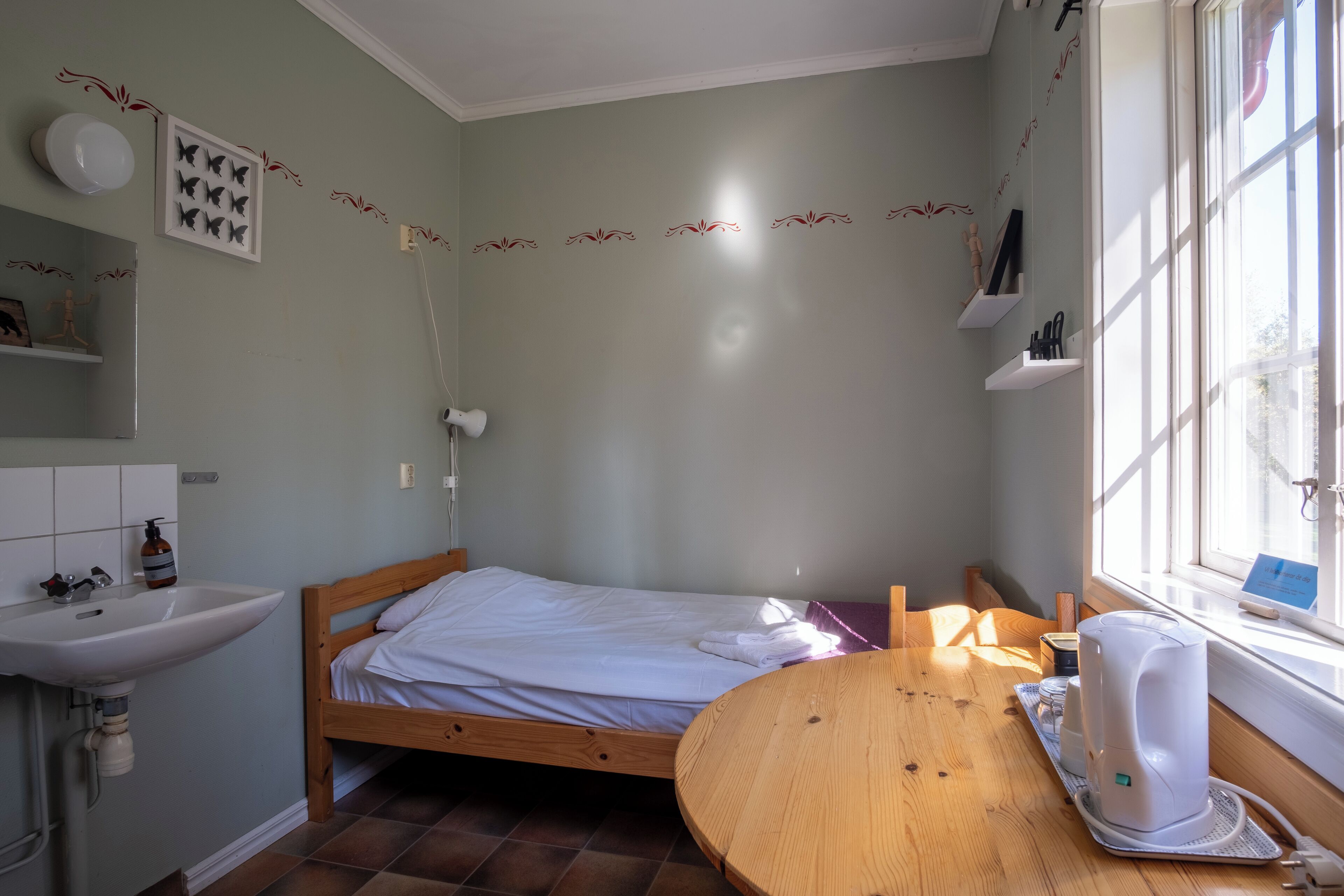 A small, well-lit room with a bed, a wooden table, a sink, and a window.