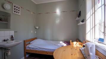 A small, well-lit room with a bed, a wooden table, a sink, and a window.