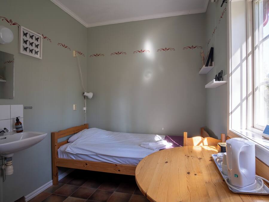 A small, well-lit room with a bed, a wooden table, a sink, and a window.