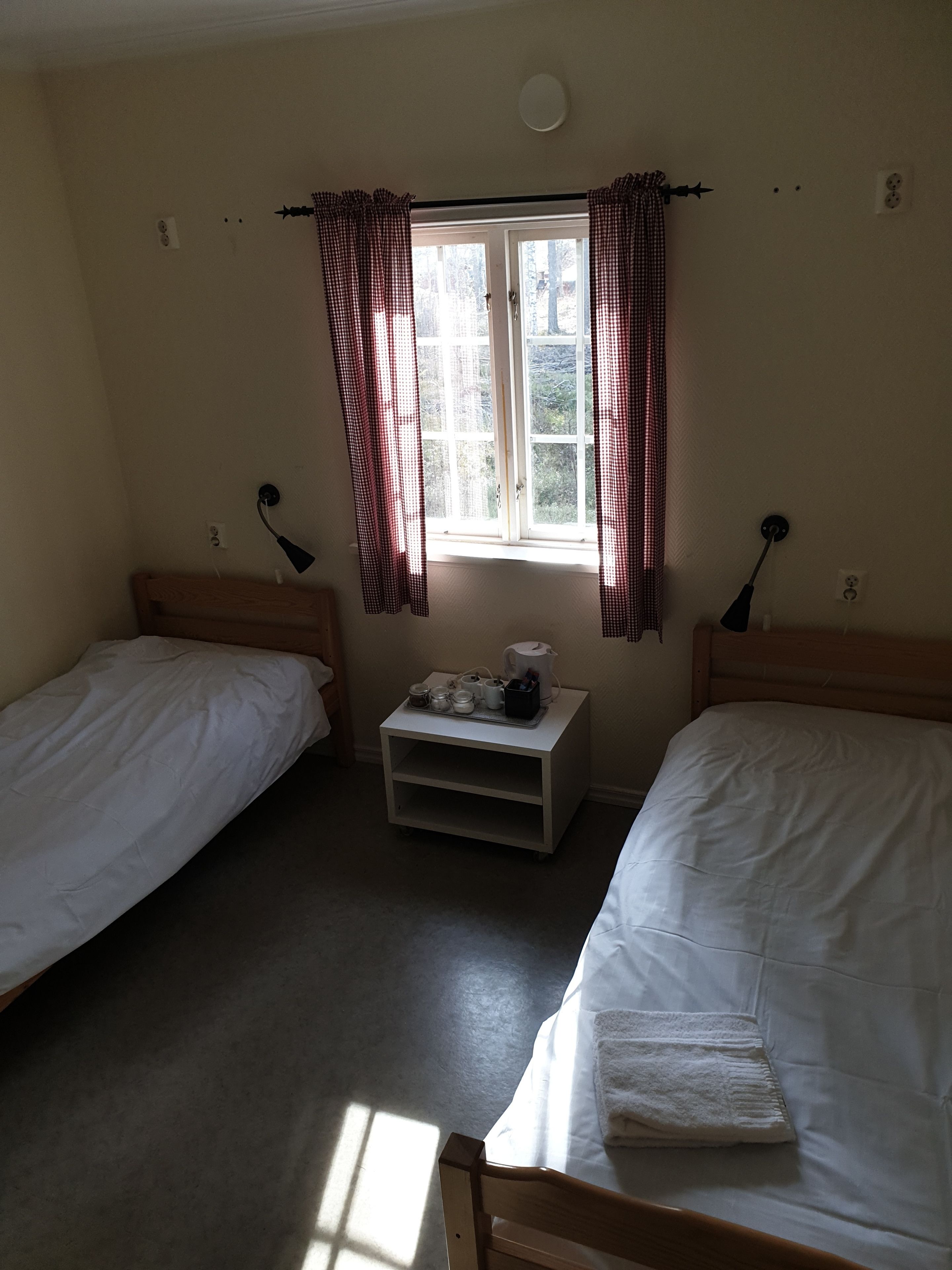 A room with two single beds, a small table with a kettle, and a window with curtains.