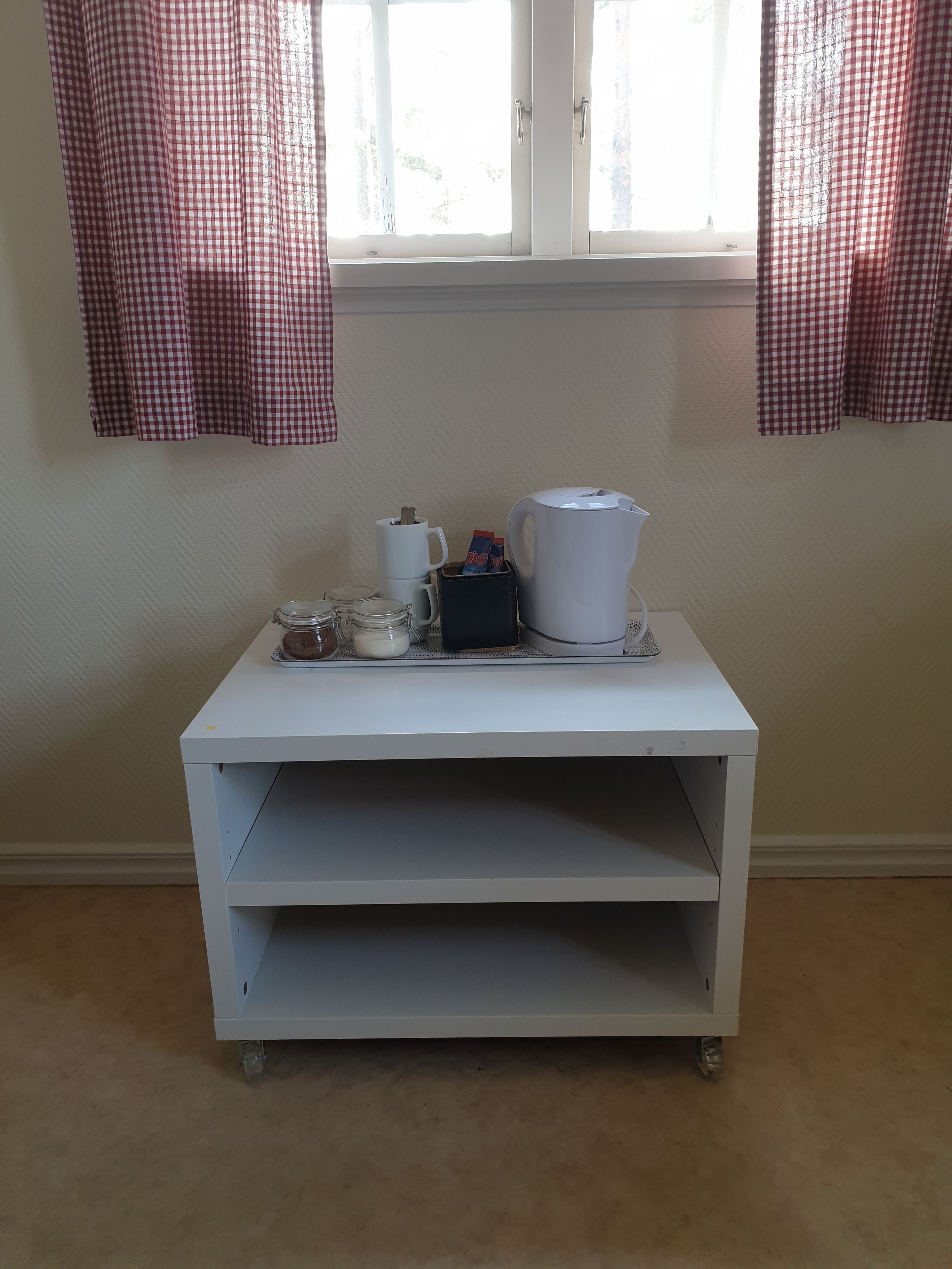 A white cart with a coffee maker, cups, and jars on a table near a window with checkered curtains.