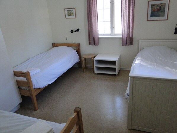 A room with two single beds, a small table, a shelf, and a window with curtains. - Sunne Hembygdsgard B&B (Sunne)