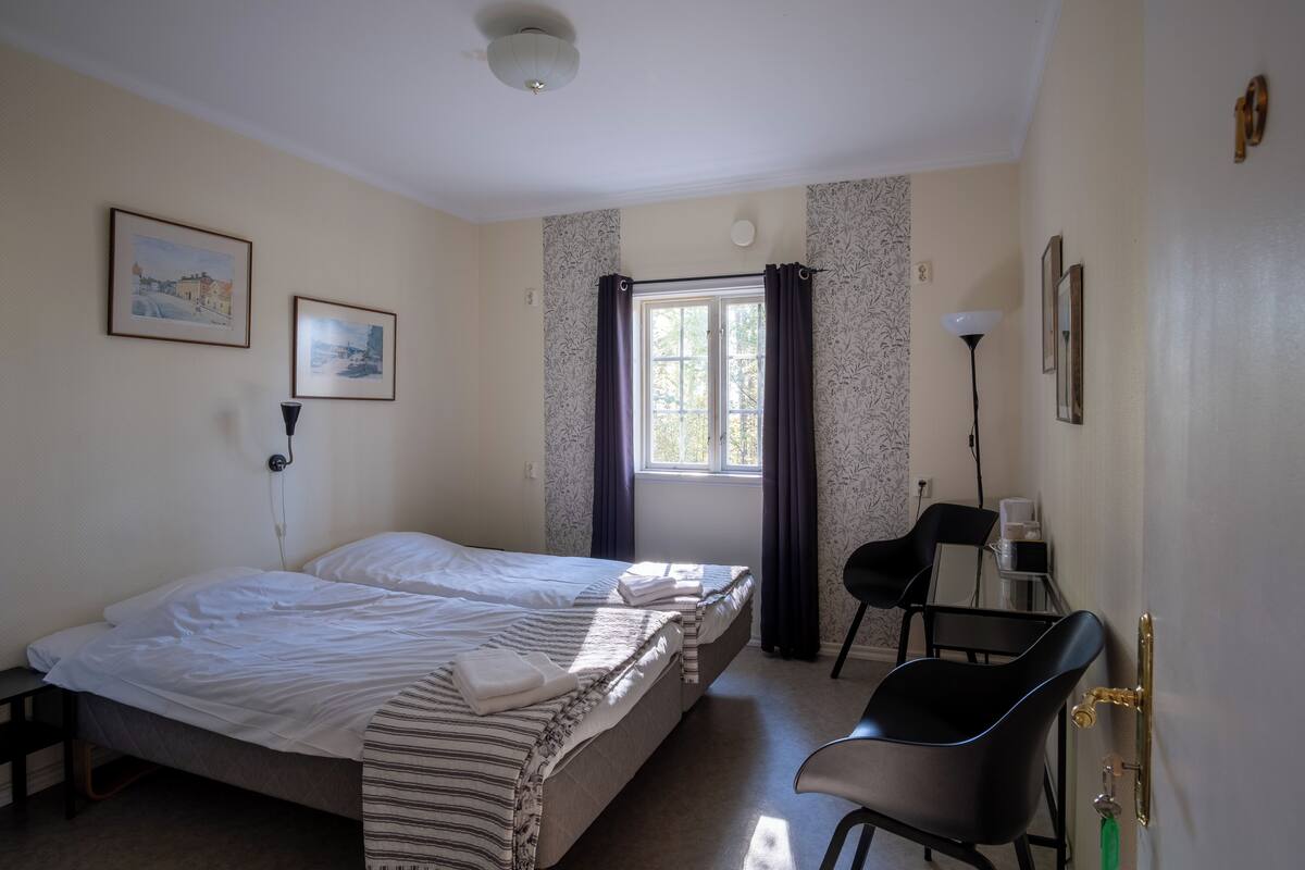 Habitación de hotel con dos camas, un escritorio, una silla, un ventanal con cortinas y obras de arte en la pared.