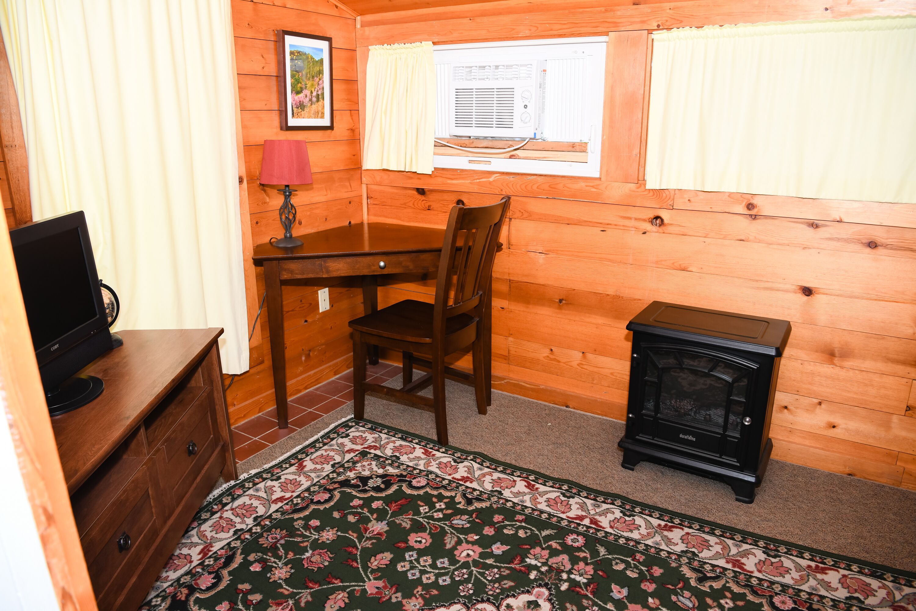 A wooden cabin room with a desk, chair, television, and a small black stove.