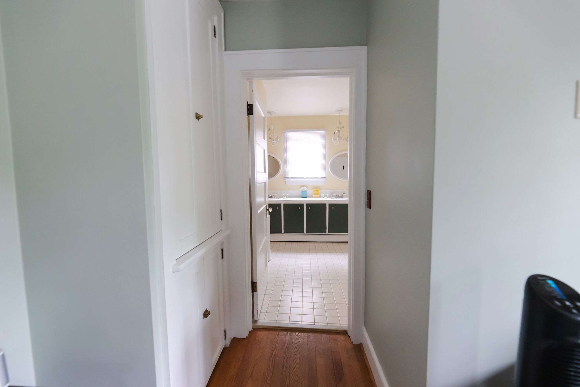 A narrow hallway leading to a kitchen with white cabinets, a window, and a tiled floor.