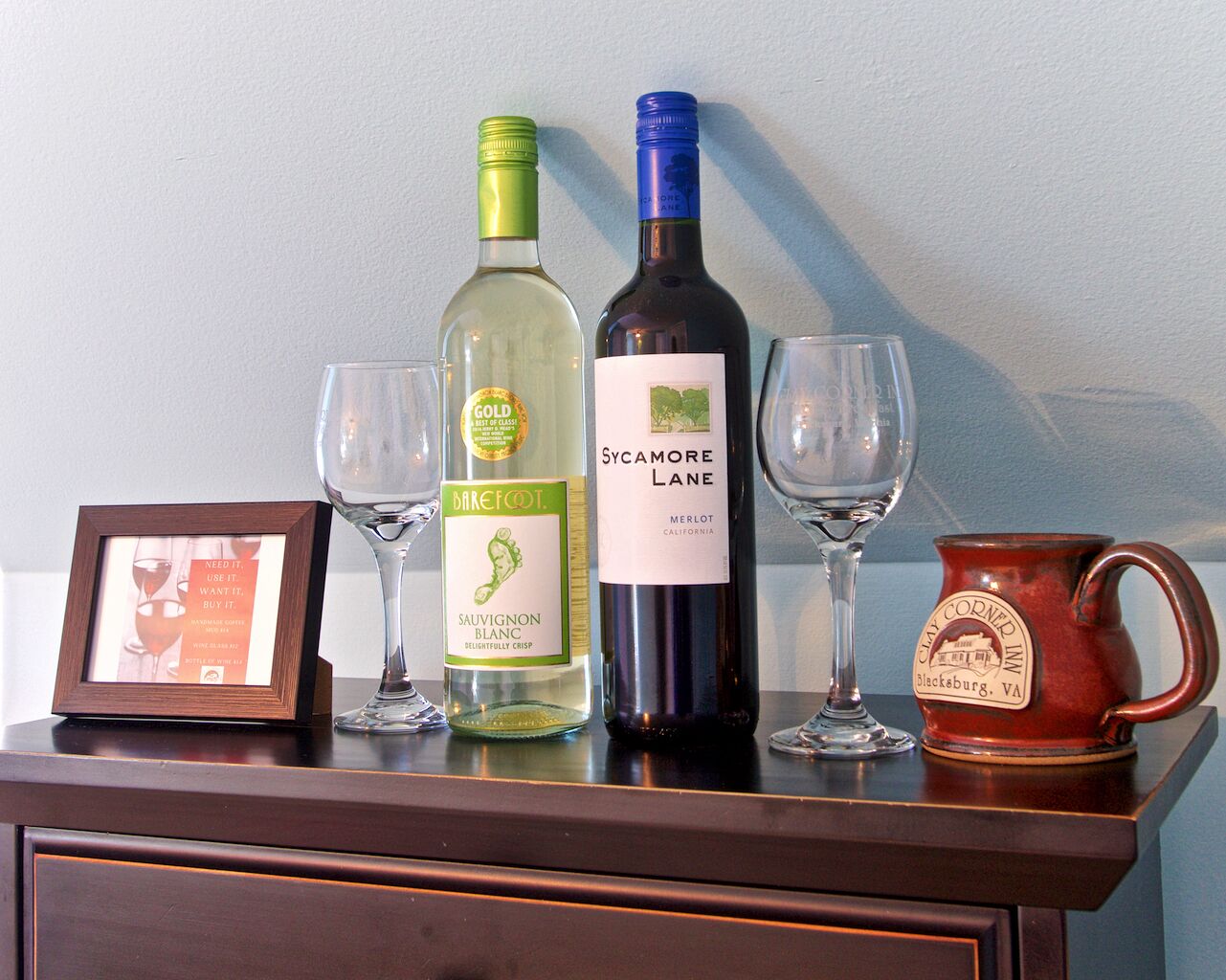 A wooden table with two wine bottles, two wine glasses, and a framed picture.