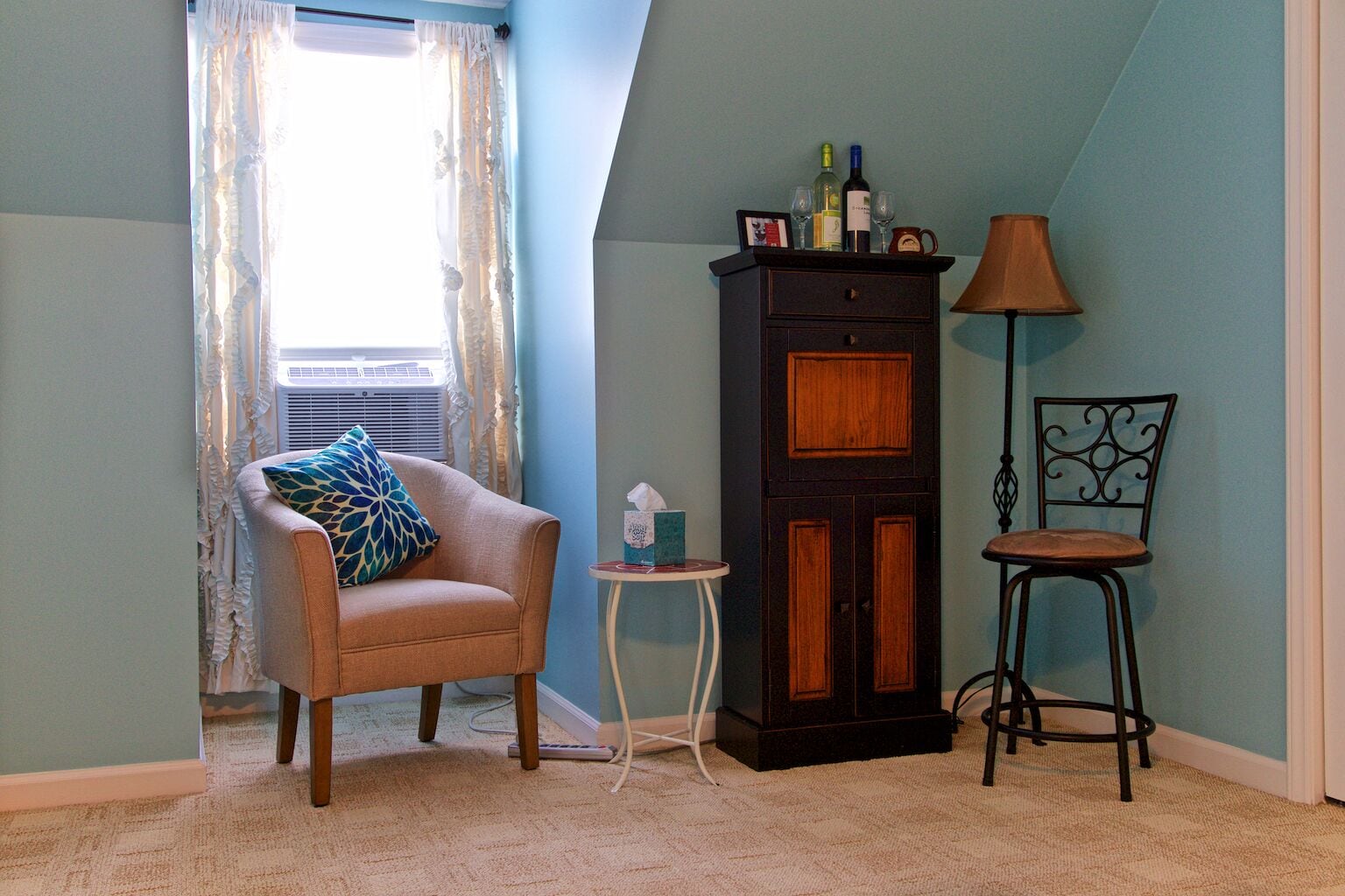 A room with a ruffled window, a cushioned armchair, a small table with a tissue box, a cabinet with bottles, a lamp, and a metal chair.