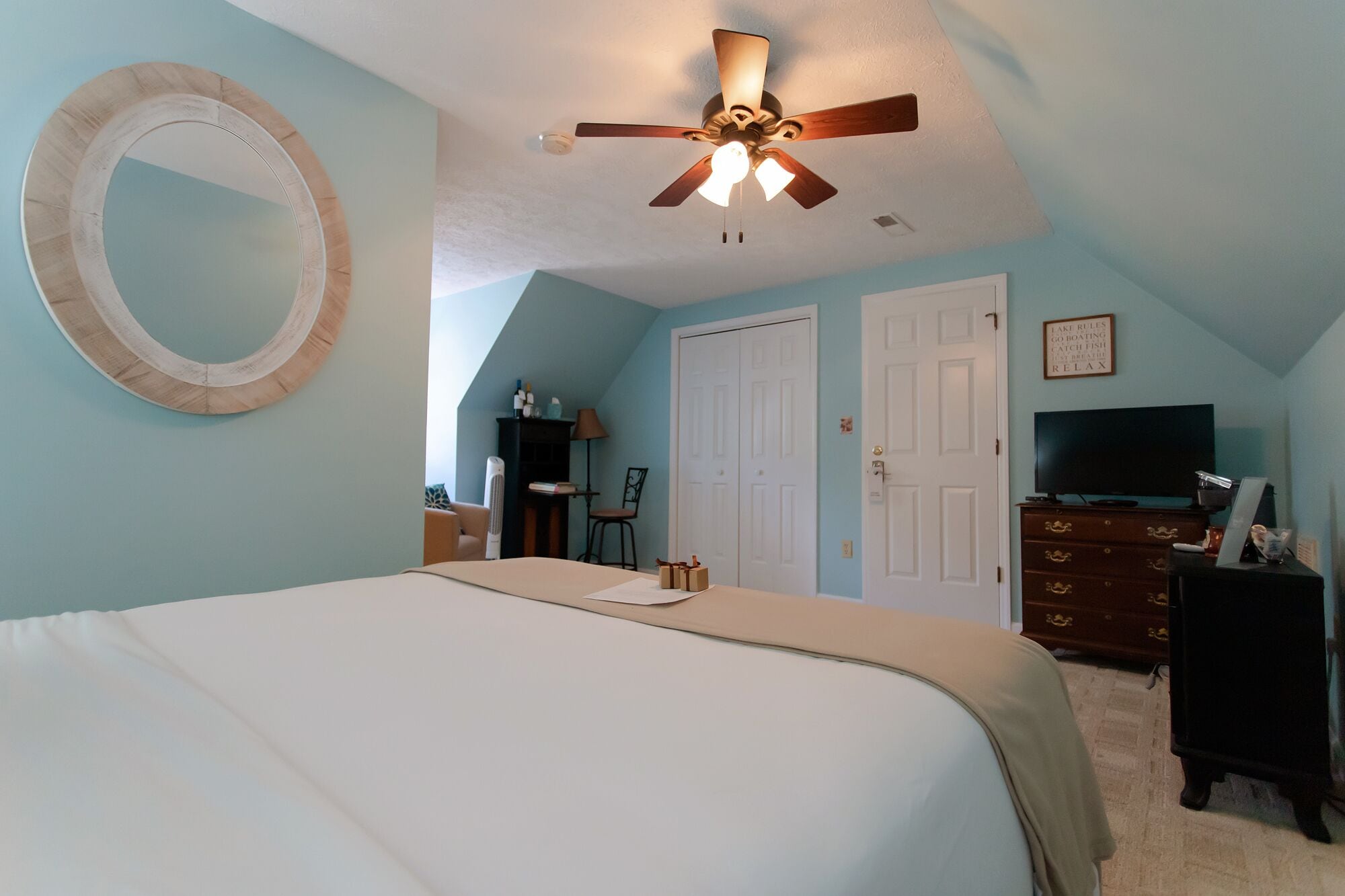 A bedroom with a bed, a ceiling fan, a mirror, a dresser, a TV, and a door.