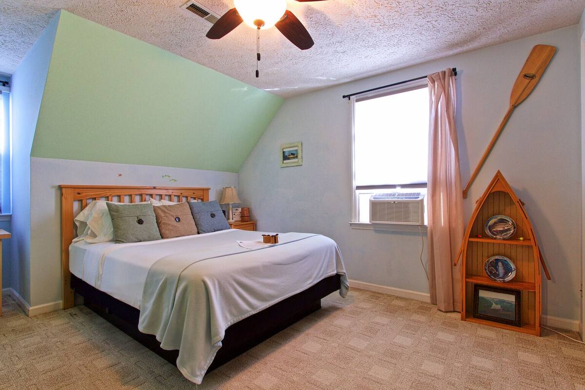 A bedroom with a bed, a ceiling fan, a window with curtains, and a small wooden shelf with decorative plates.