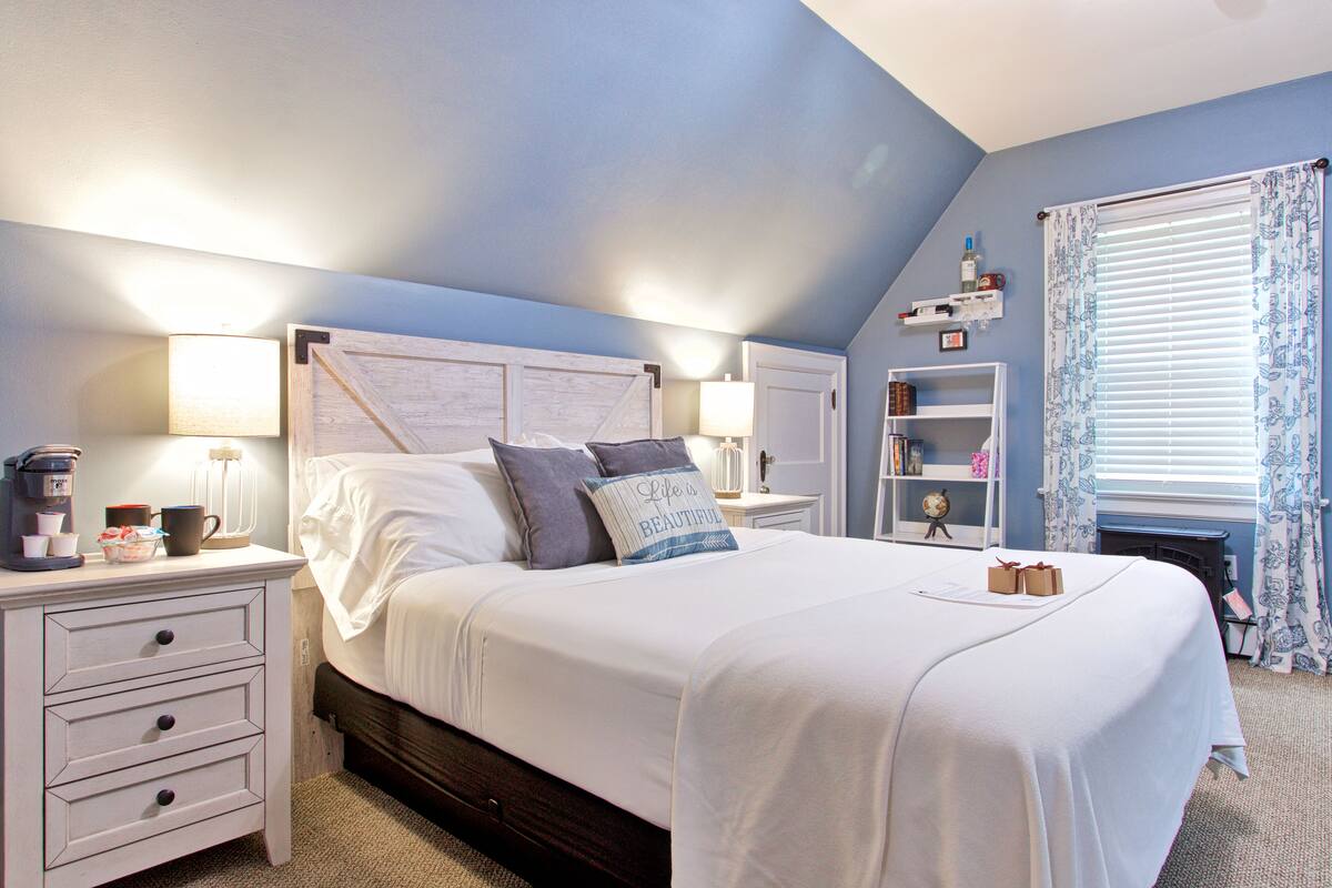 A bedroom with a large bed, two bedside tables, a nightstand, a lamp, a shelf with books and a clock, and a window with curtains.