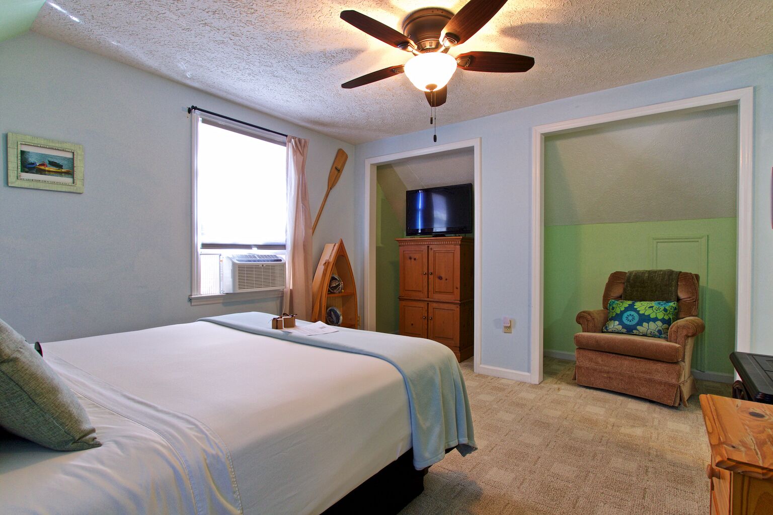 A bedroom with a bed, a ceiling fan, a television, a chair, and a window with curtains.