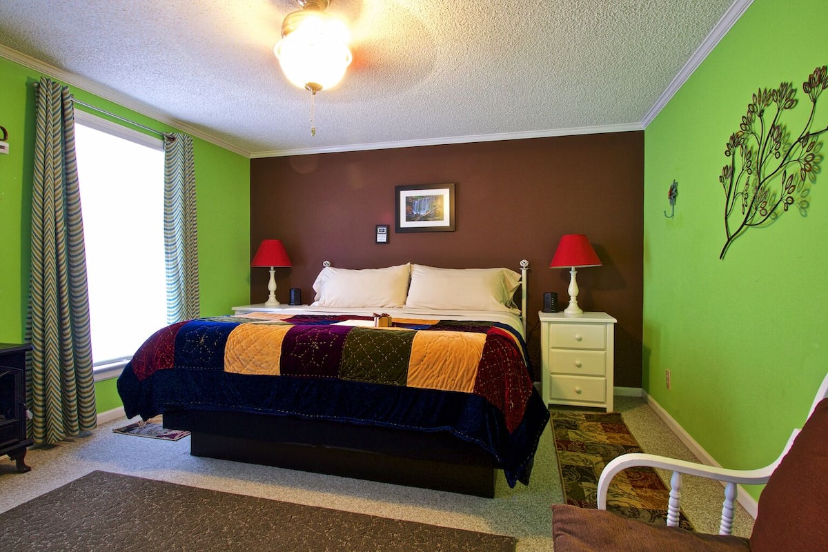 A bedroom with a bed, two bedside tables, a nightstand, a ceiling fan, a window with curtains, and a wall decoration.