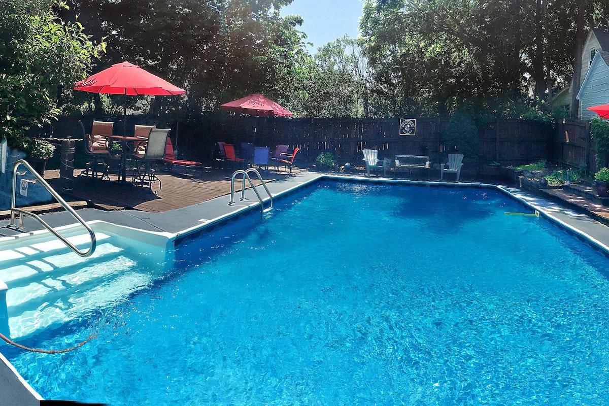 A rectangular swimming pool with a ladder on one side, surrounded by a wooden deck. There are red umbrellas and outdoor furniture, including chairs and a table.