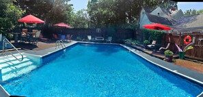 A rectangular swimming pool with a ladder on one side, surrounded by a wooden deck. There are red umbrellas and outdoor furniture, including chairs and a table.
