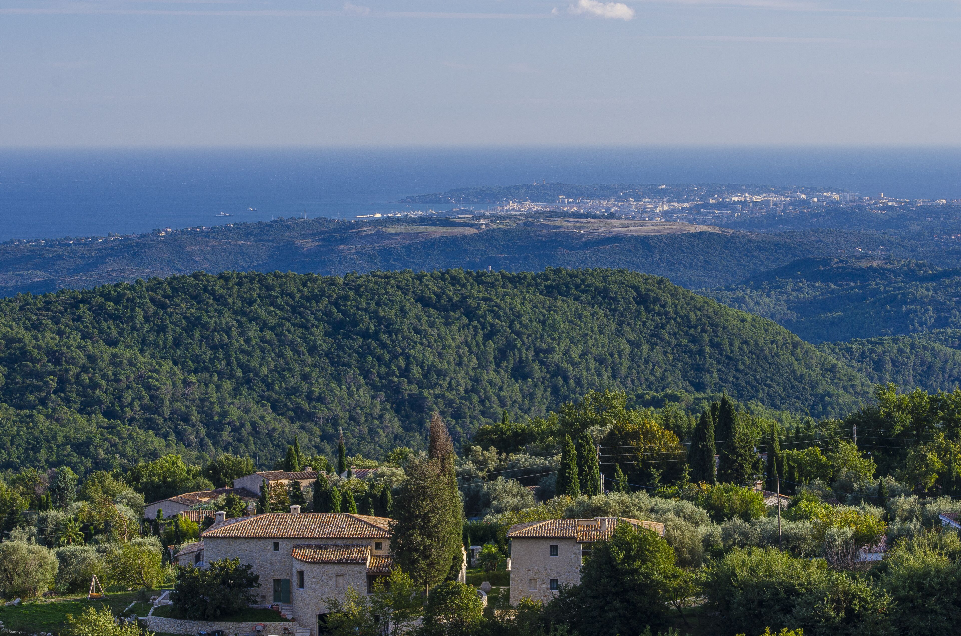 A rural village with stone houses, dense greenery, and a view of the sea and distant city.