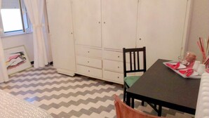 A room with a chevron-patterned floor, a white wardrobe, a black table with a red placemat, and a chair with a green cushion.