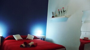 A neatly made bed with red bedding, a small bedside table with a lamp, and a wall-mounted shelf with toiletries.