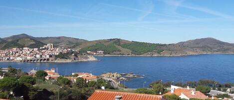 A coastal town with terracotta-roofed buildings, a bridge, and hills in the background.