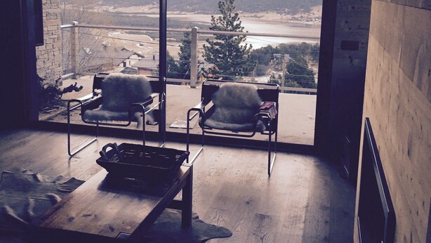 A room with a large window overlooking a scenic view of mountains and a river. There are two chairs and a table in the room. - Large chalet 12 people (Les Angles)