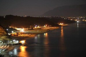 A nighttime view of a coastal area with a river, illuminated buildings, and a distant mountain range.