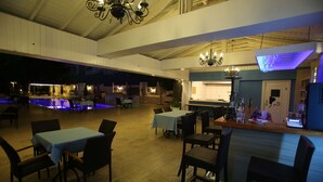 Un amplio comedor interior con varias mesas y sillas, un área de piscina con iluminación azul, y un bar con distintos artículos en la barra.