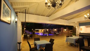 Un área de comedor al aire libre con mesas y sillas, una piscina al fondo y un techo con candelabros.