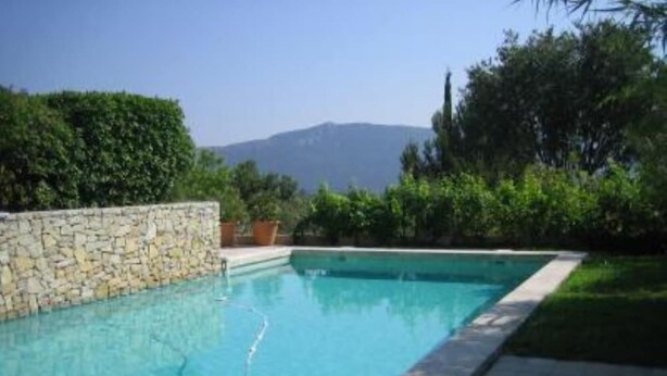 A rectangular swimming pool with a stone wall on one side, surrounded by greenery and a clear sky.