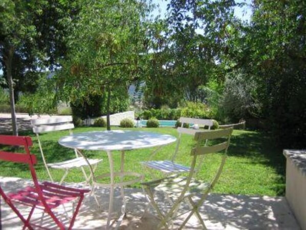 Un patio al aire libre con una mesa redonda y cuatro sillas, rodeado de exuberante vegetación y una piscina al fondo.