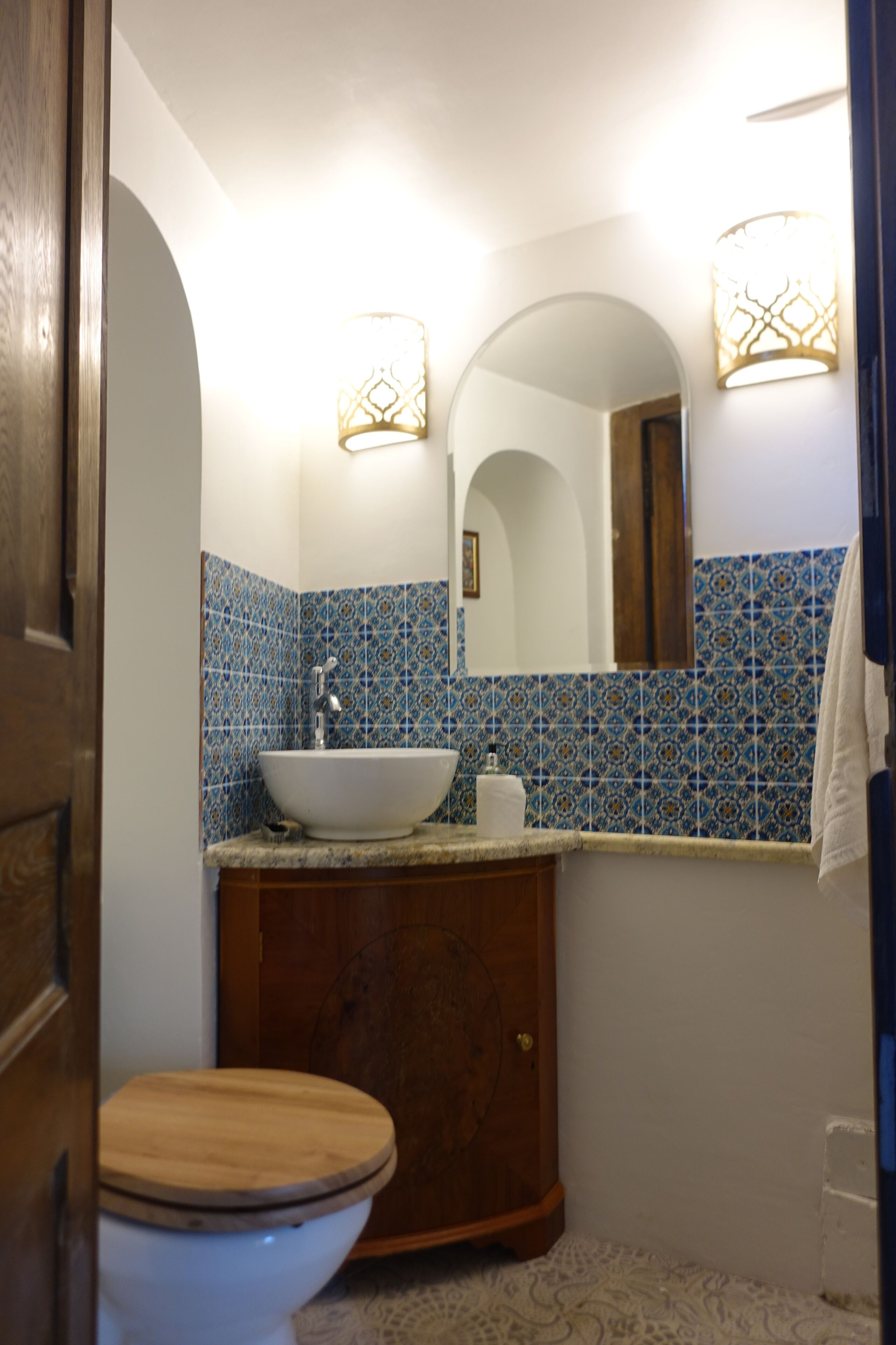 A bathroom with a wooden toilet, a white sink, and a mirror above it. There is a towel hanging on the wall and a patterned tile backsplash.