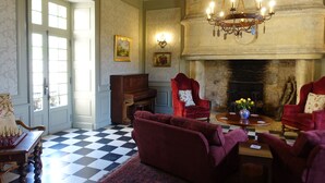 A room with a checkered floor, a fireplace, red armchairs, a wooden piano, and a chandelier.