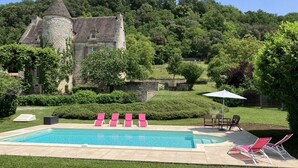 A swimming pool with pink lounge chairs, a stone building with a tower, and a lush green landscape.