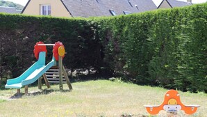 Un parque infantil con tobogán y estructura para escalar, rodeado de un seto y una casa al fondo.