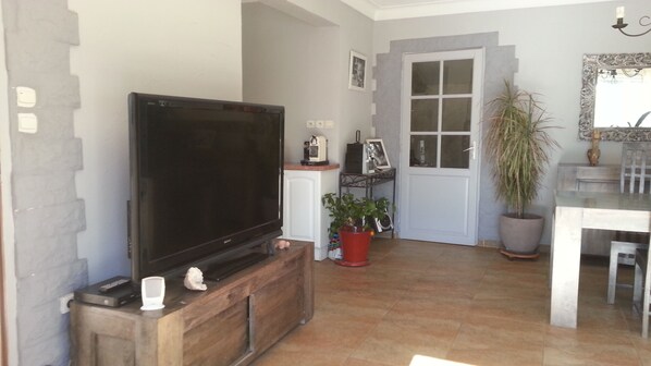 Una sala de estar con televisor de pantalla plana sobre un mueble de madera, una planta en maceta, una mesa de comedor con sillas y una puerta blanca con paneles de vidrio.