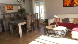 Un comedor con una mesa de madera y sillas, una mesa redonda de centro con base metálica, un sofá blanco con cojines y un espejo grande en la pared.