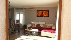 Salón con sofá blanco, manta roja y un cuadro de flores. Hay una mesita redonda con una planta y una silla con respaldo de listones de madera.